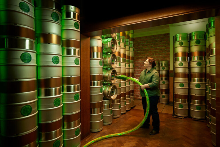 Worker plugging a glowing green fibre cable into stacked 'Fast Streaming Internet' kegs in a green-lit cellar