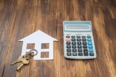 A paper house cut-out with house keys beside a calculator on a wooden surface, symbolising home loan or mortgage costs