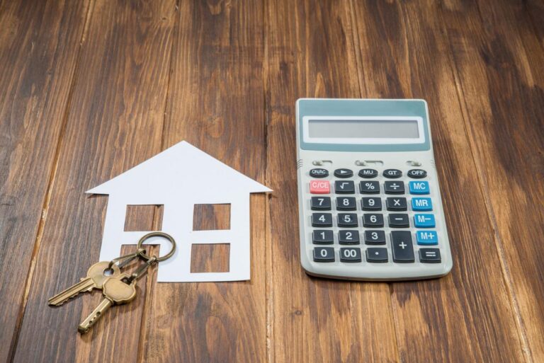 A paper house cut-out with house keys beside a calculator on a wooden surface, symbolising home loan or mortgage costs