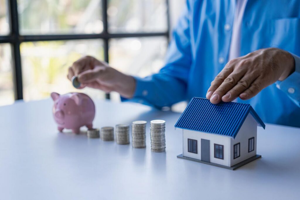 Hand placing a coin in a piggy bank next to coin stacks and a model house