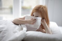 A little girl lying on a bed, sneezing with a tissue in her hand.