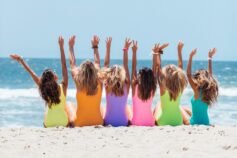 A group of women in colourful swimsuits sitting together on a beach with their arms raised