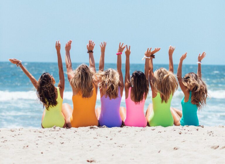 A group of women in colourful swimsuits sitting together on a beach with their arms raised