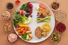 Overhead view of a white plate divided into vegetables, fruit, wholegrains and protein foods, surrounded by small bowls of nuts, spices and pink salt