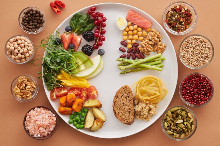 Overhead view of a white plate divided into vegetables, fruit, wholegrains and protein foods, surrounded by small bowls of nuts, spices and pink salt