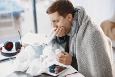 Sick man wrapped in a blanket sitting at a table with cold medicine, tissues and a warm drink.