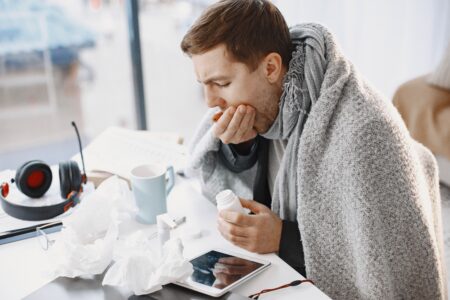 Sick man wrapped in a blanket sitting at a table with cold medicine, tissues and a warm drink.