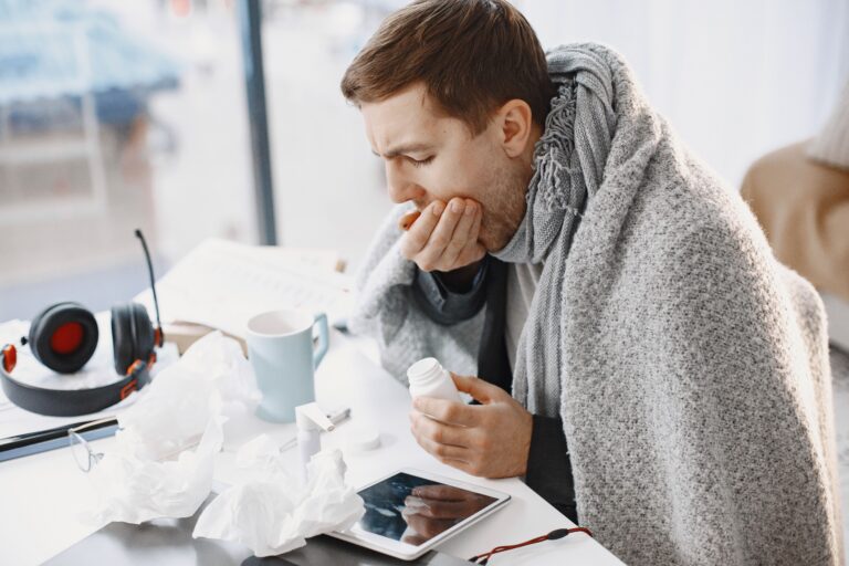 Sick man wrapped in a blanket sitting at a table with cold medicine, tissues and a warm drink.